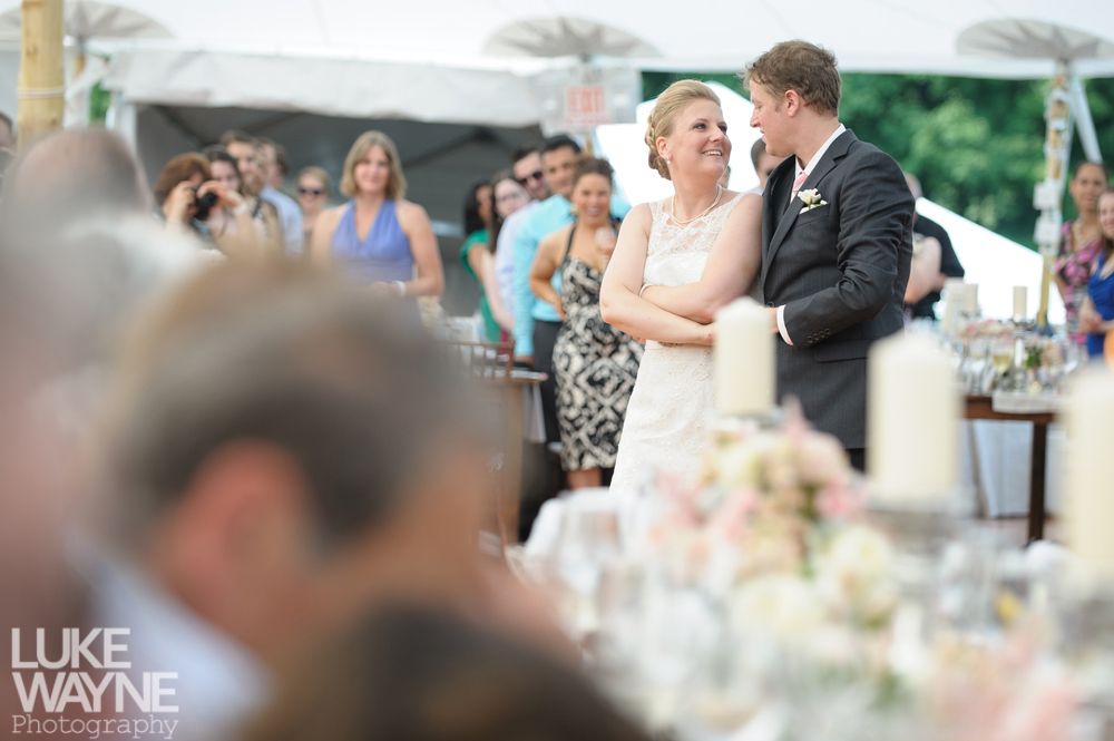Bride and groom at wedding reception, smiling at each other. Guests and tent visible in the background.
