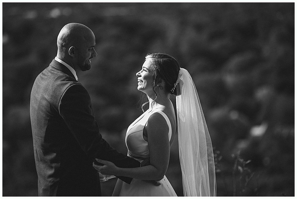 Wedding ceremony. Guests seated, smiling. Groom facing officiant. Bride in white dress. Outdoors, sunny day.