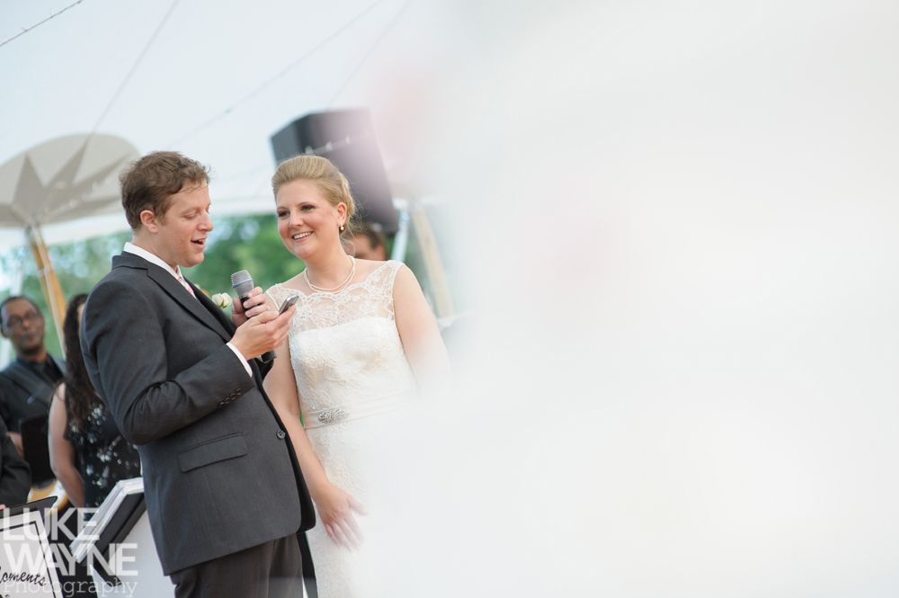 Groom speaking into a microphone, looking at smiling bride in a wedding dress, outdoor ceremony.