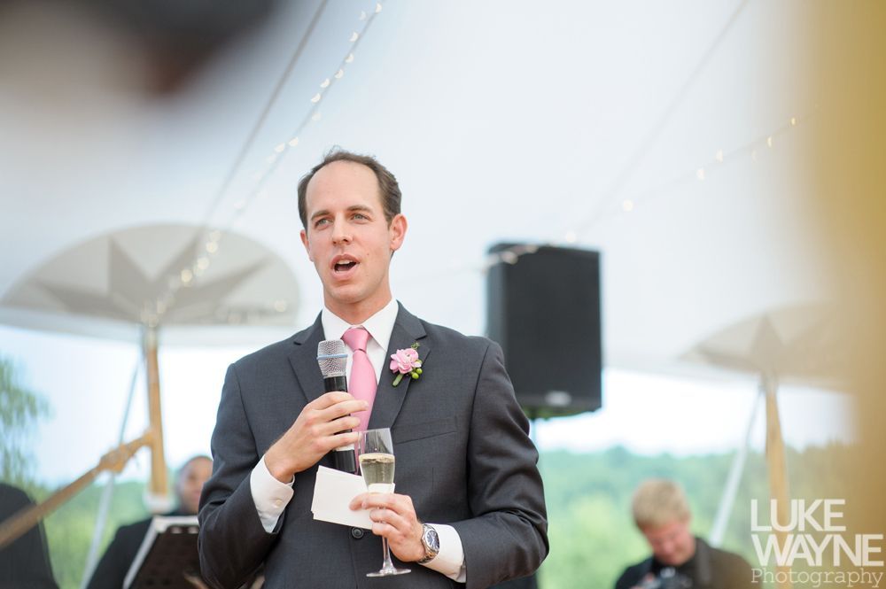 Man in gray suit giving a speech at an outdoor wedding, holding a microphone and champagne.