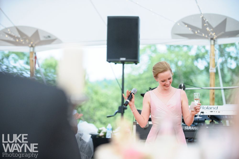 Woman in pink dress giving a speech at a wedding, holding a microphone and champagne. Outdoor setting with a tent.