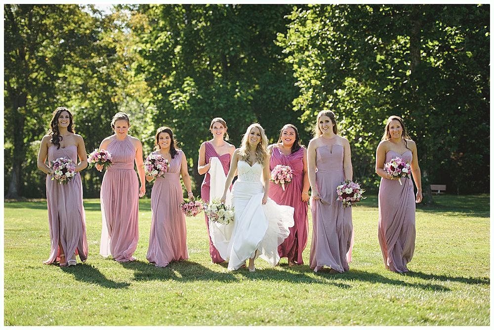 Bride smiles at the altar during a ceremony. Groomsmen in suits partially block view. Outdoor setting.