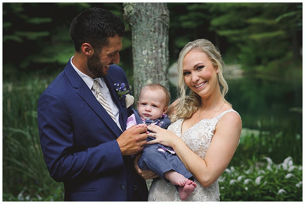 Wedding ceremony outdoors by a lake with a couple, wedding party, and guests under a cloudy sky.