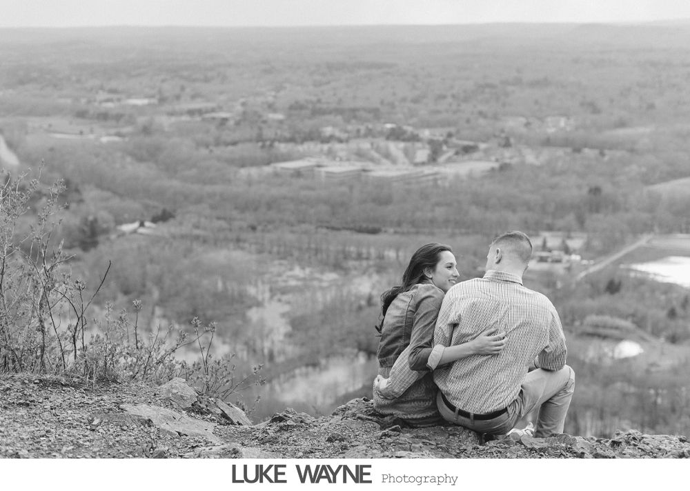 Couple embraces, overlooking a scenic valley. Woman looks toward man; both are seated. Black and white photo.