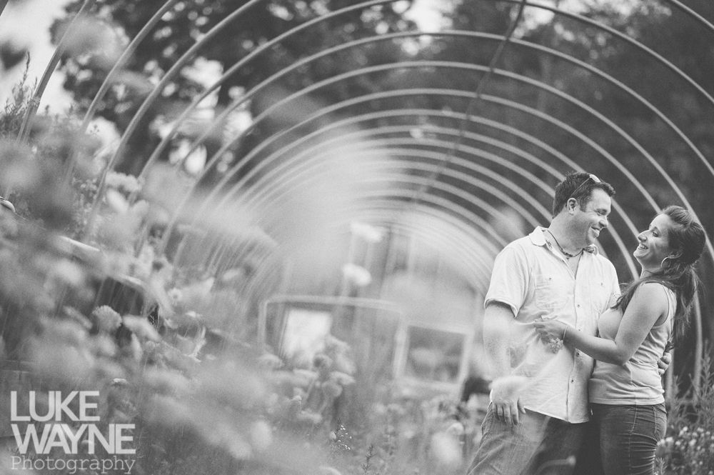 Couple smiling, holding hands, under metal archway. Plants on either side, black and white photograph.