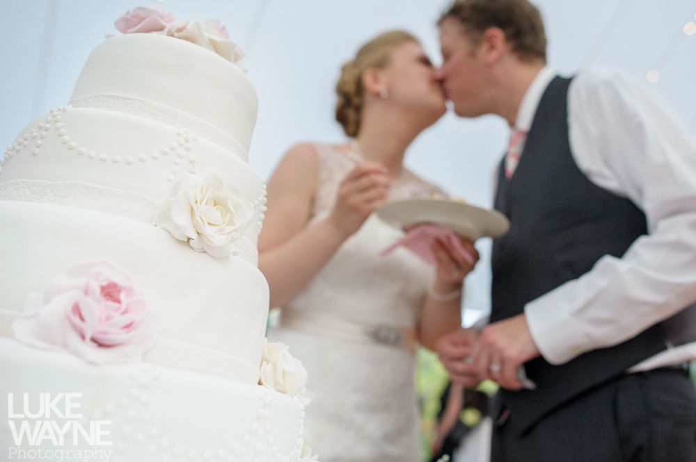 Wedding cake in focus; couple blurred in background, kissing, outdoors.