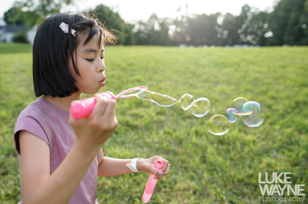 Girl blowing bubbles in a grassy field. She holds a pink bubble wand and wears a pink shirt.