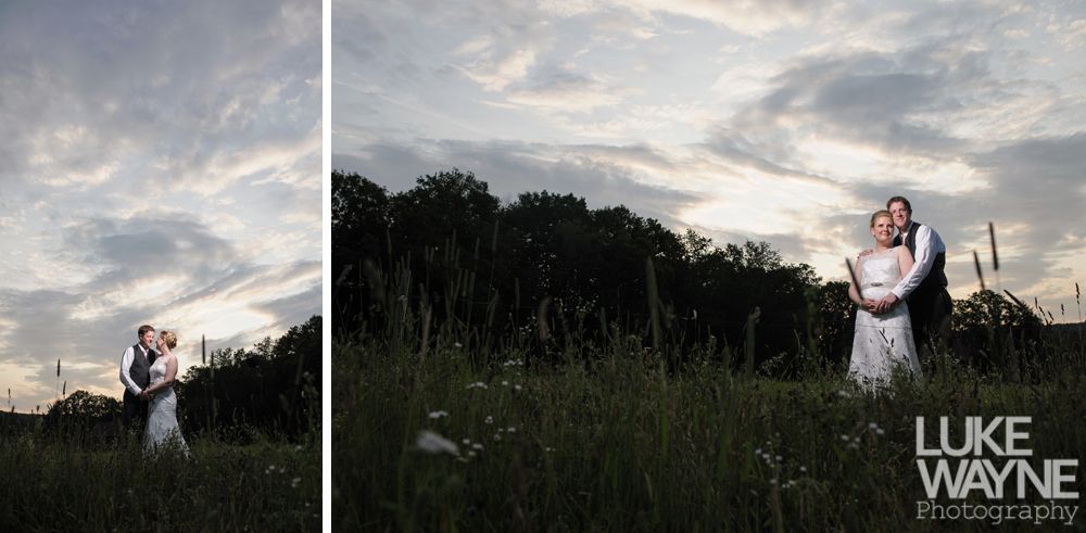 Couple embracing in a field during sunset, wearing wedding attire. Dark trees in background, tall grass in foreground.
