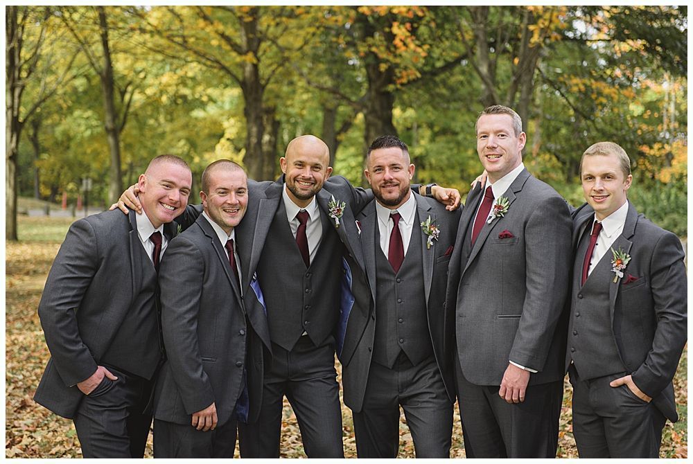 Wedding party poses outdoors. Groomsmen in navy suits, bridesmaids in red dresses, bride in white dress, holding bouquets.