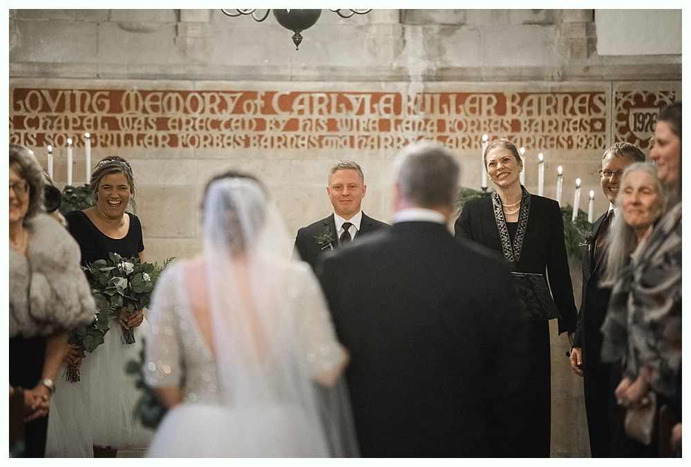 Groom kissing bride's cheek; she holds flowers. They are outdoors, near a tree. She wears a veil and dress, he wears a suit.