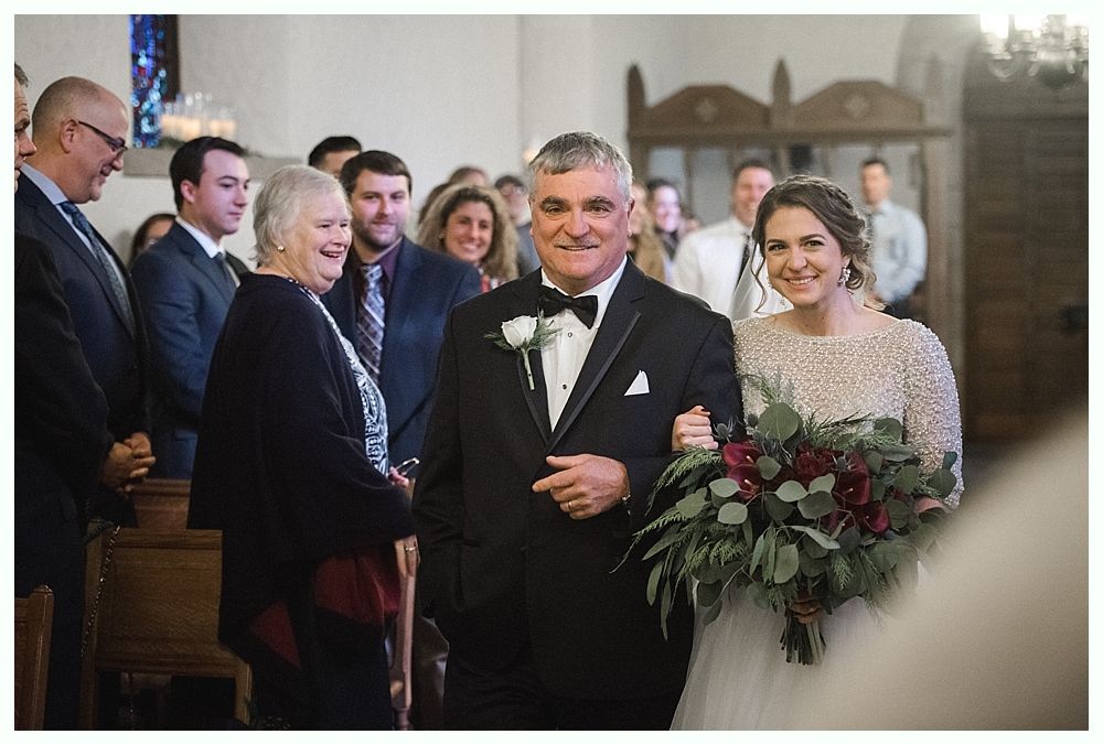 Bride and groom embracing, smiling, holding flowers. Trees and water in the background. Black and white.