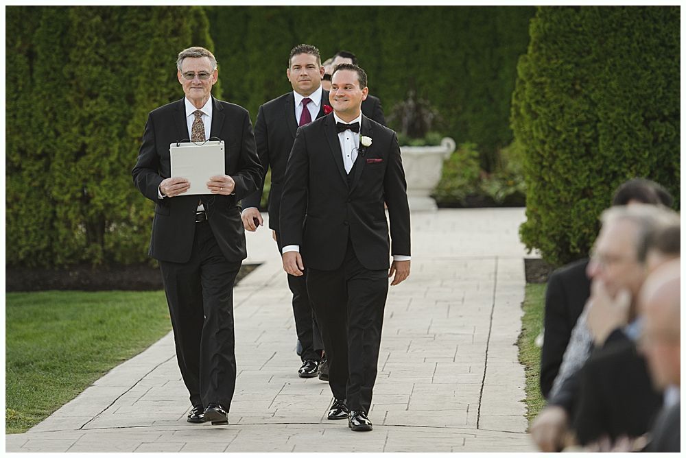 Groom kissing bride's cheek; she holds flowers. They are outdoors, near a tree. She wears a veil and dress, he wears a suit.