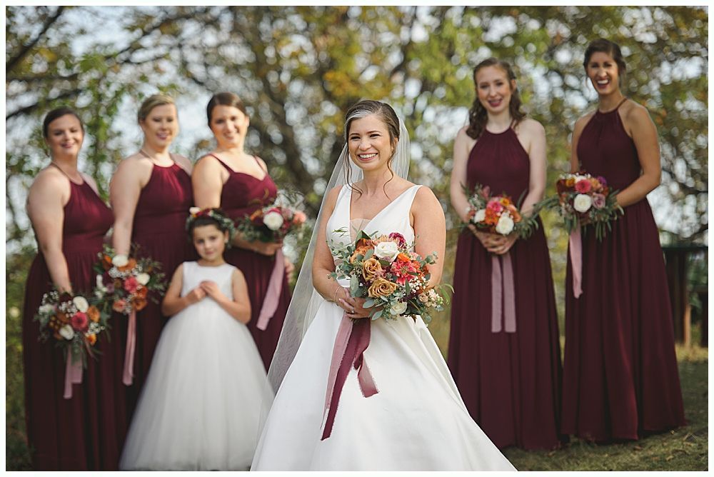 Wedding party smiling outdoors near water. Bride and bridesmaids in red, groom and groomsmen in navy.