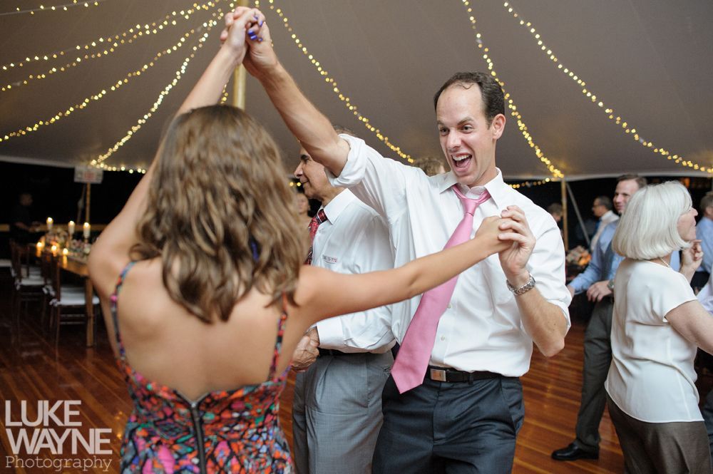 People dancing under a tent at a party, man in pink tie leading a woman, smiling, with overhead lights.