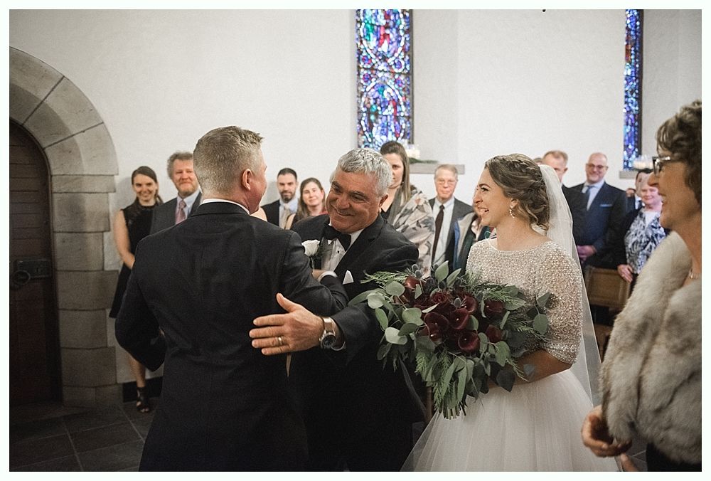 Bride and groom walk hand-in-hand across a grassy hilltop; black and white photograph with a cloudy sky backdrop.