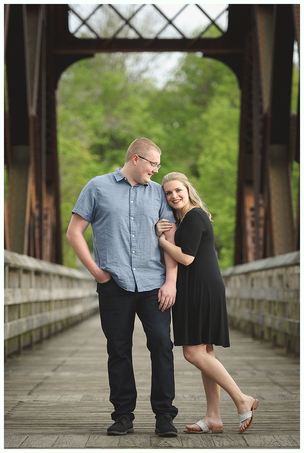Couple on a wooden bridge, man in blue shirt and jeans, woman in black dress, smiling. Green trees background.