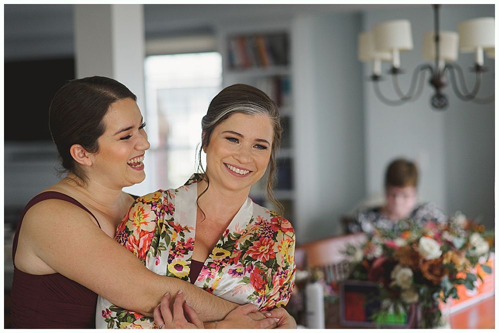Group of women in white robes, sitting on a couch, smiling and laughing. One woman is being held by others.