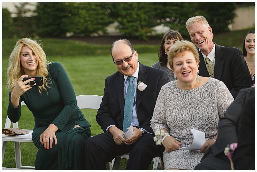 Bride and groom embracing, smiling, holding flowers. Trees and water in the background. Black and white.