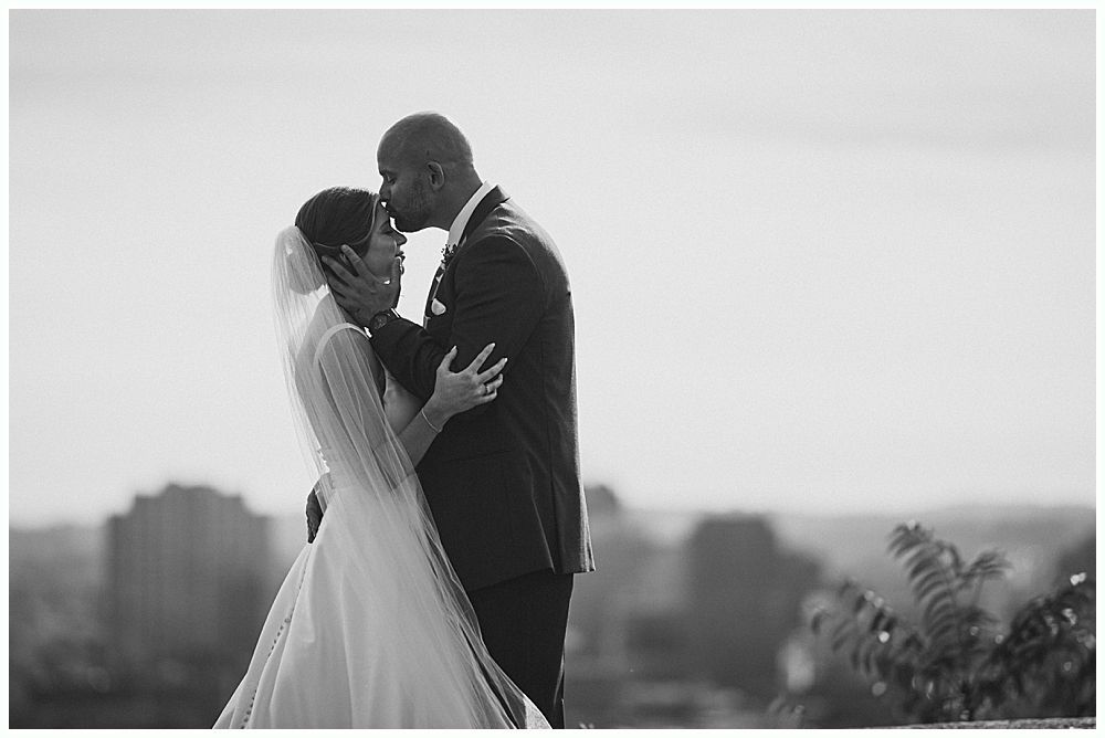 Groom kissing bride's cheek; she holds flowers. They are outdoors, near a tree. She wears a veil and dress, he wears a suit.