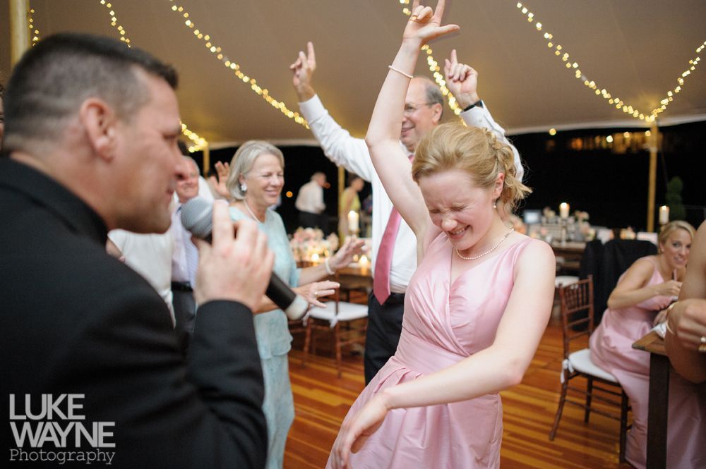 DJ singing at a wedding reception; a woman in a pink dress dances joyfully, others celebrate.