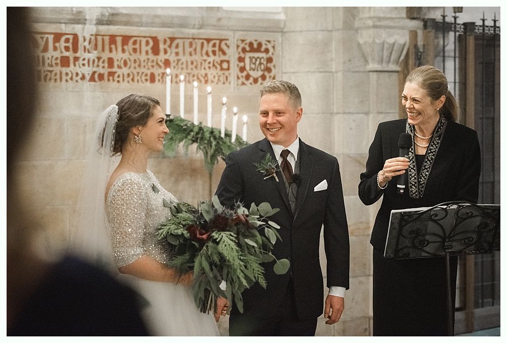 Bride touches groom's face; he holds a bouquet. They smile outdoors.