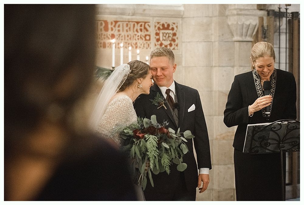 Bride and groom holding hands, smiling. She wears a white dress and veil, he wears a blue suit. Outdoors, near trees.