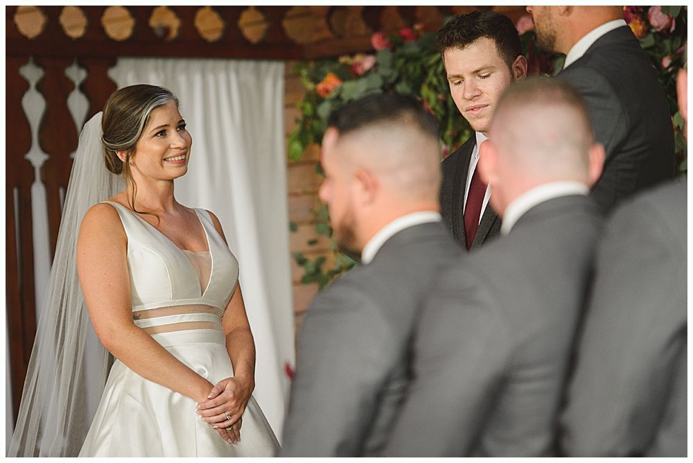 Bride touches groom's face; he holds a bouquet. They smile outdoors.