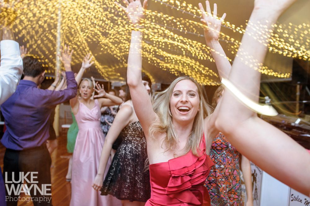 People dancing at a party with arms raised. Woman in red dress smiles. String lights hang above.