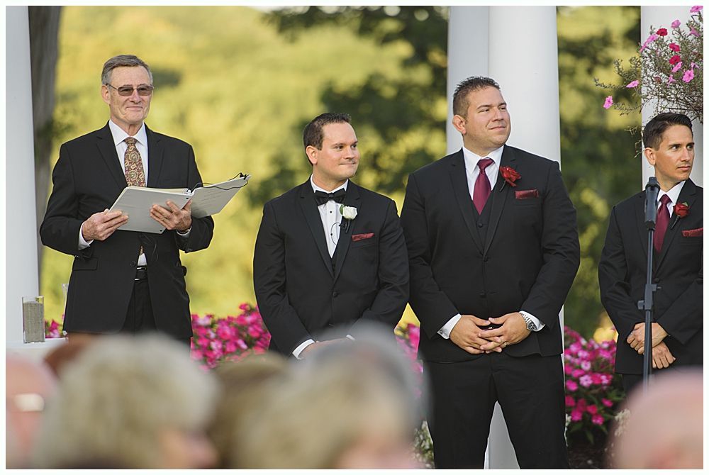 Bride and groom holding hands, smiling. She wears a white dress and veil, he wears a blue suit. Outdoors, near trees.