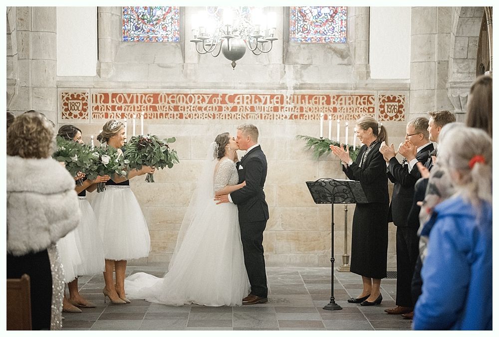 Bride and groom pose near a tree and pond. Bride in lace dress holds bouquet. Groom embraces her from behind.