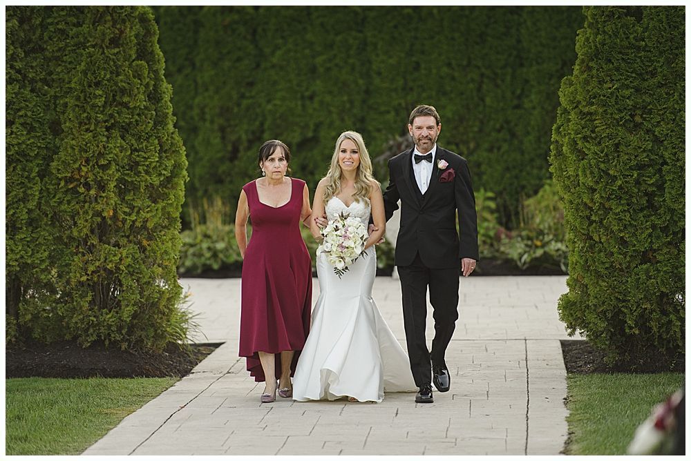 Couple in wedding attire embrace under a large weeping willow tree on a grassy hill.