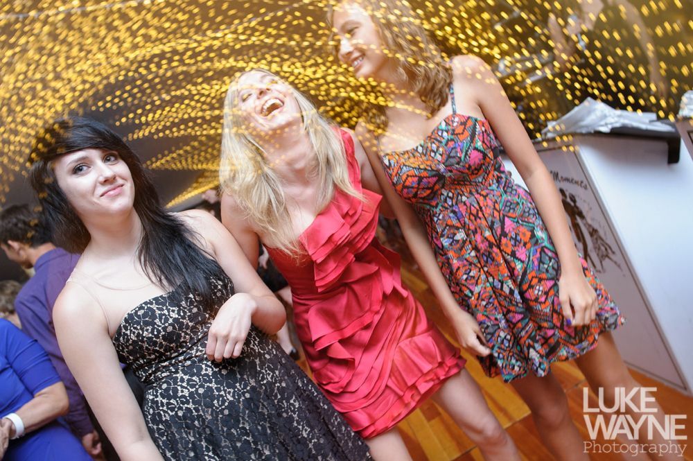 Three women in party dresses laugh. One in red, one in patterned, one in black with a backdrop of golden lights.