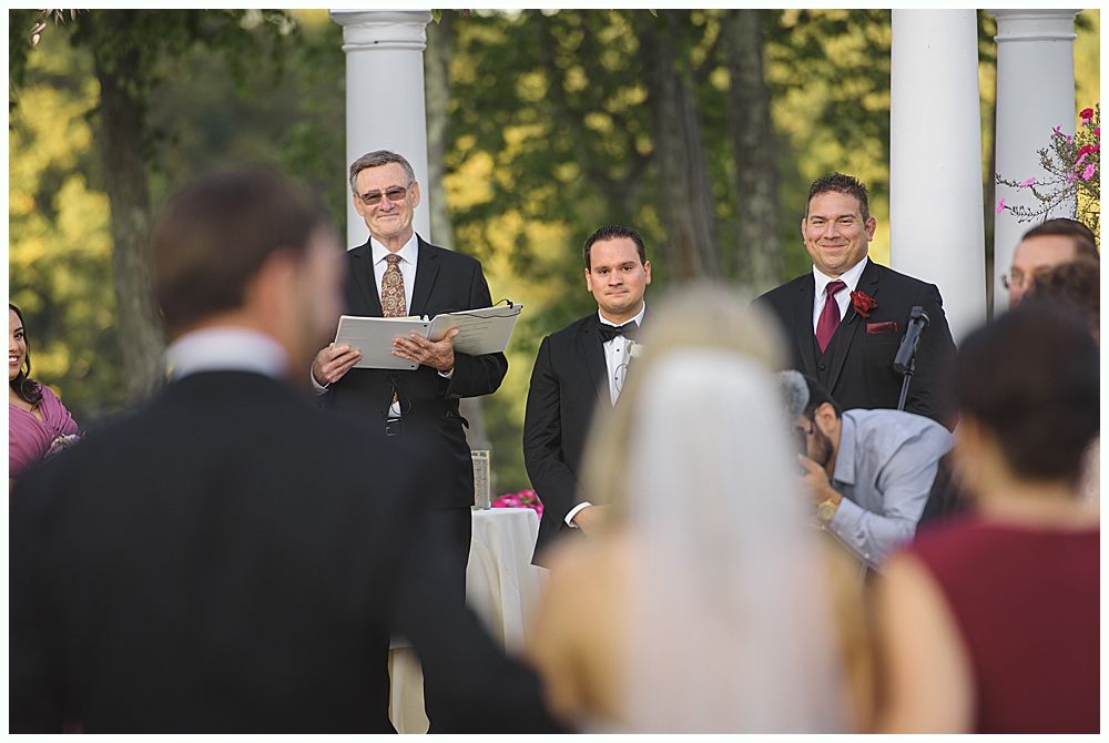 Bride and groom pose near a tree and pond. Bride in lace dress holds bouquet. Groom embraces her from behind.