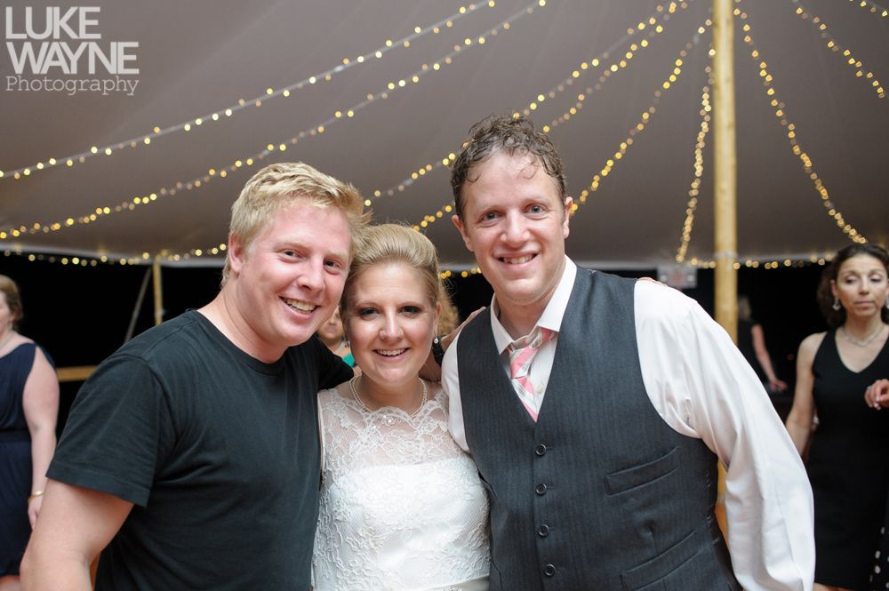 Three people smile at the camera under a tent with string lights; one wears a wedding dress, one a black tee, and one a vest.