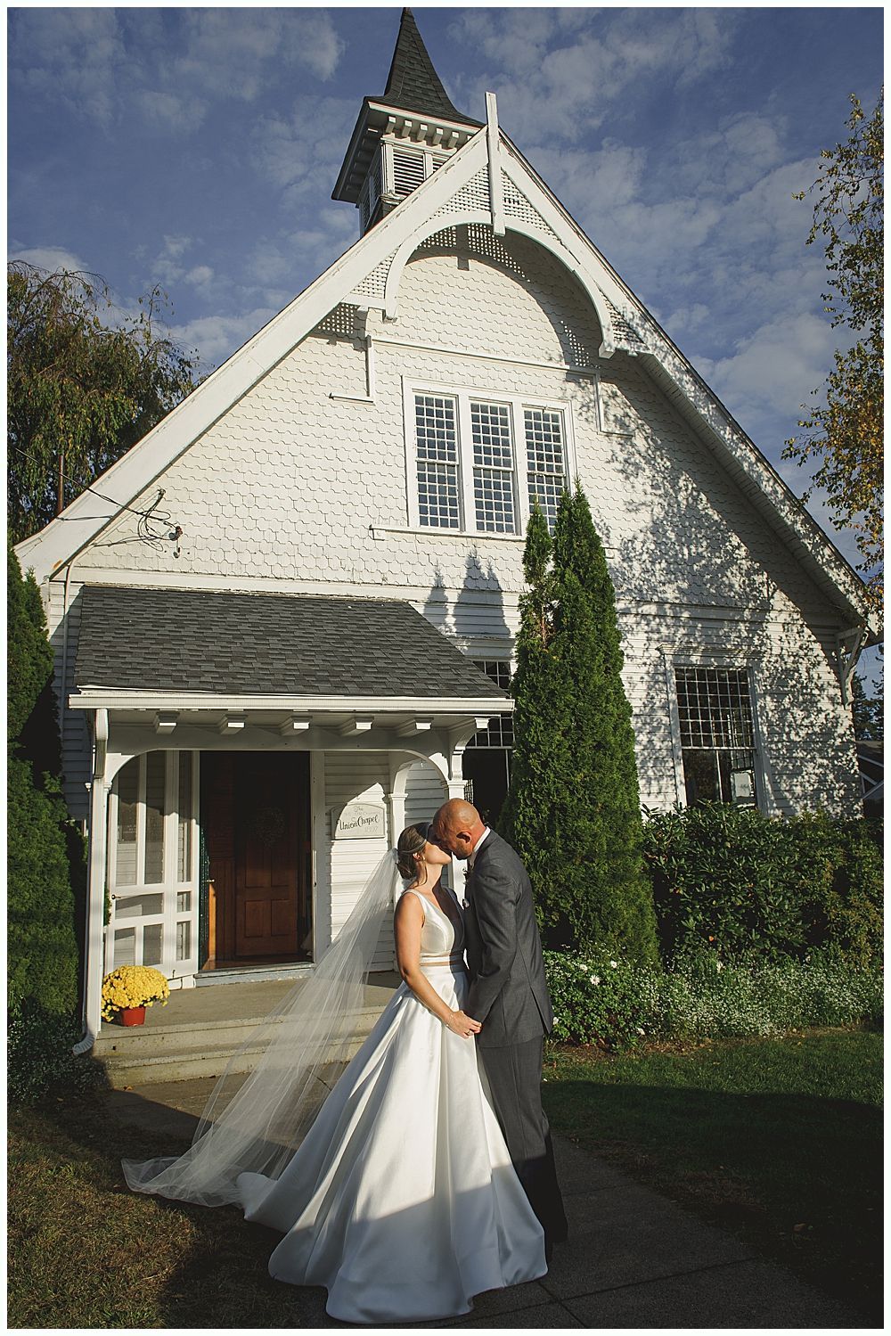Bride and groom pose near a tree and pond. Bride in lace dress holds bouquet. Groom embraces her from behind.