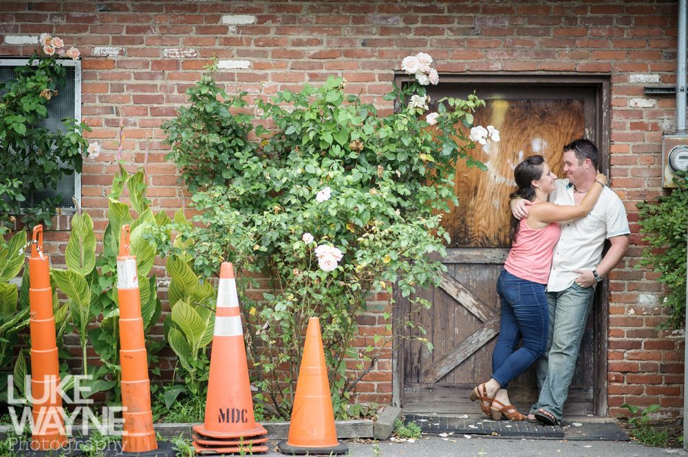 Couple embracing by a brick building with greenery, orange traffic cones in foreground.
