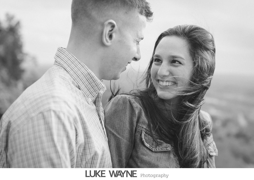 Couple gazing at each other and smiling; woman's hair blowing; outdoor setting with blurry background.