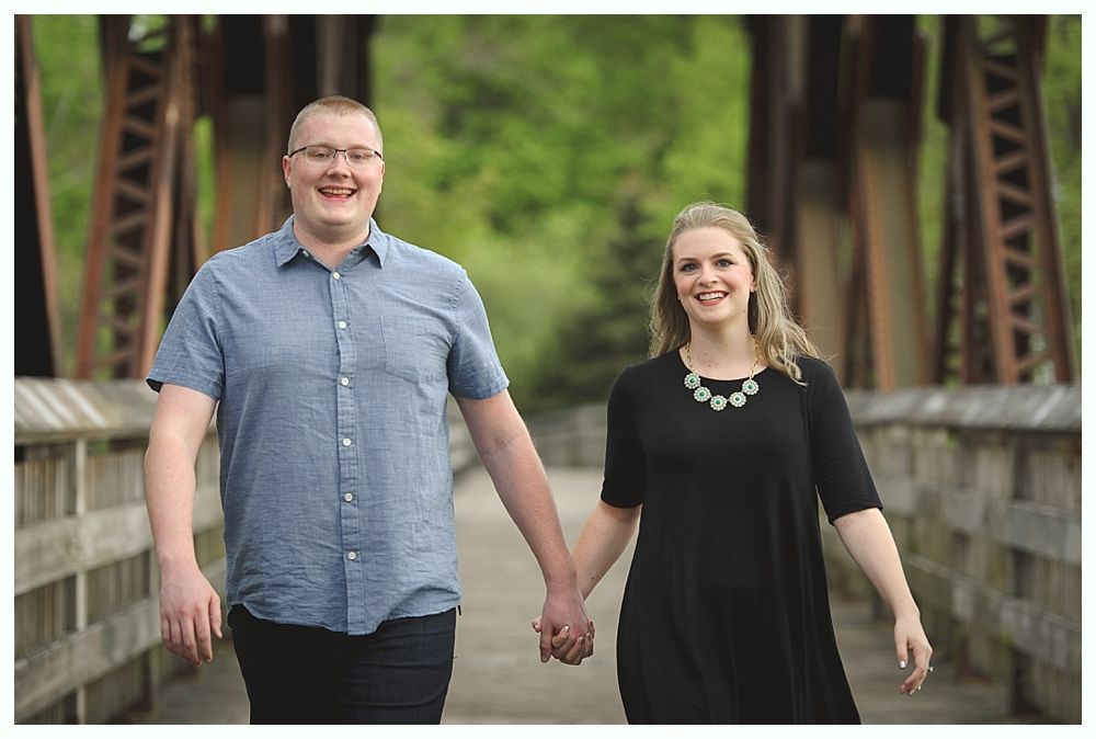 Couple holding hands, smiling while walking on a wooden bridge, wearing casual clothing, nature background.