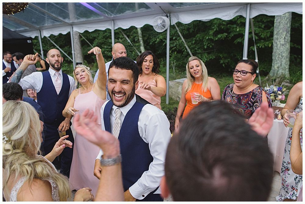A man in a suit gives a speech at a wedding. The bride and groom laugh while listening.