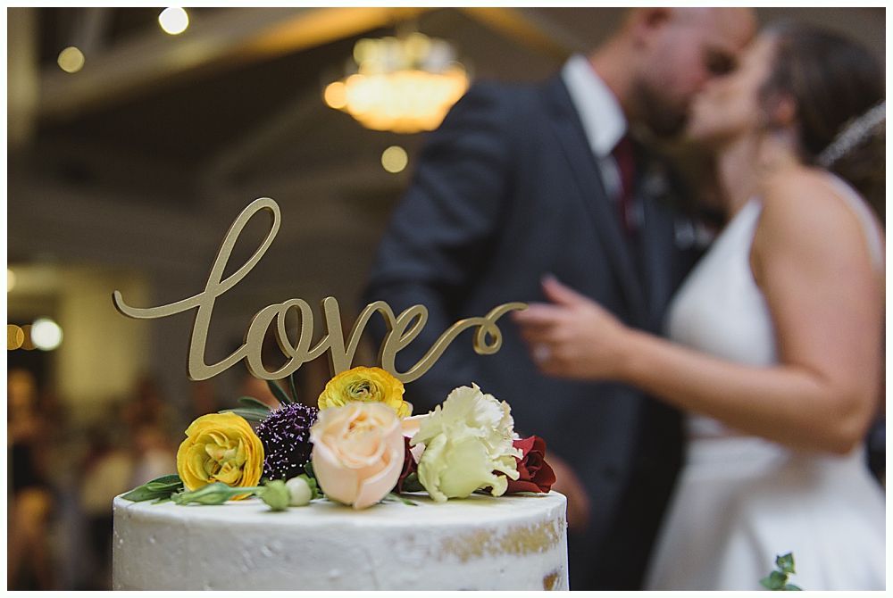 A man in a suit gives a speech at a wedding. The bride and groom laugh while listening.