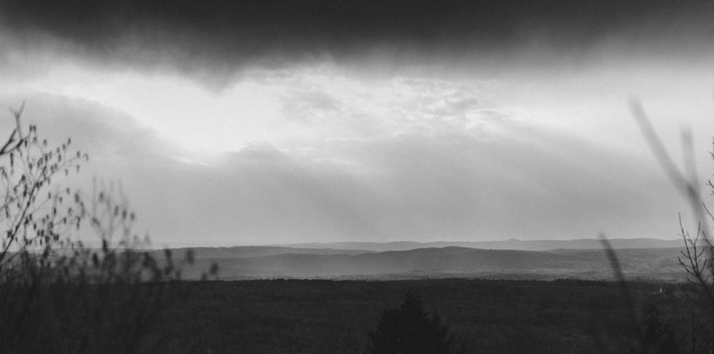 Black and white landscape with a dark, cloudy sky over a field. Sunbeams filter through the clouds.
