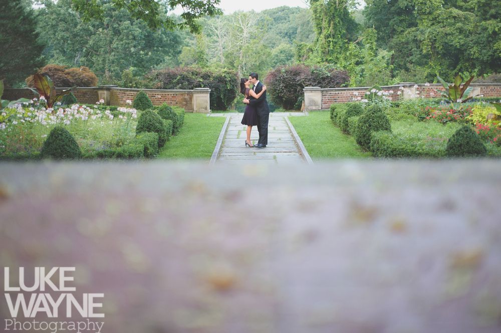 Couple embracing in a garden, standing on a stone path, framed by hedges and flowers.