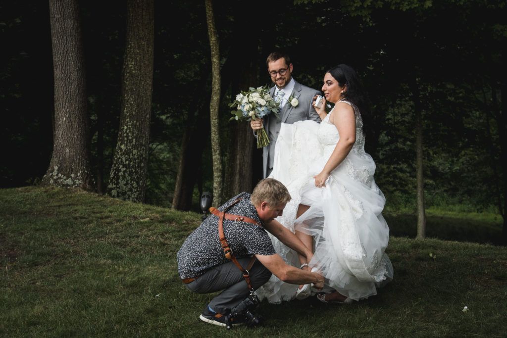 Photographer adjusts bride's shoe as she poses with groom, outdoors.