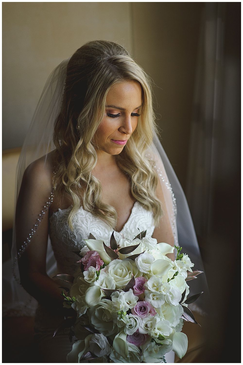 Bride holding bouquet; burgundy, cream, and green flowers. She looks down, lit by soft light.