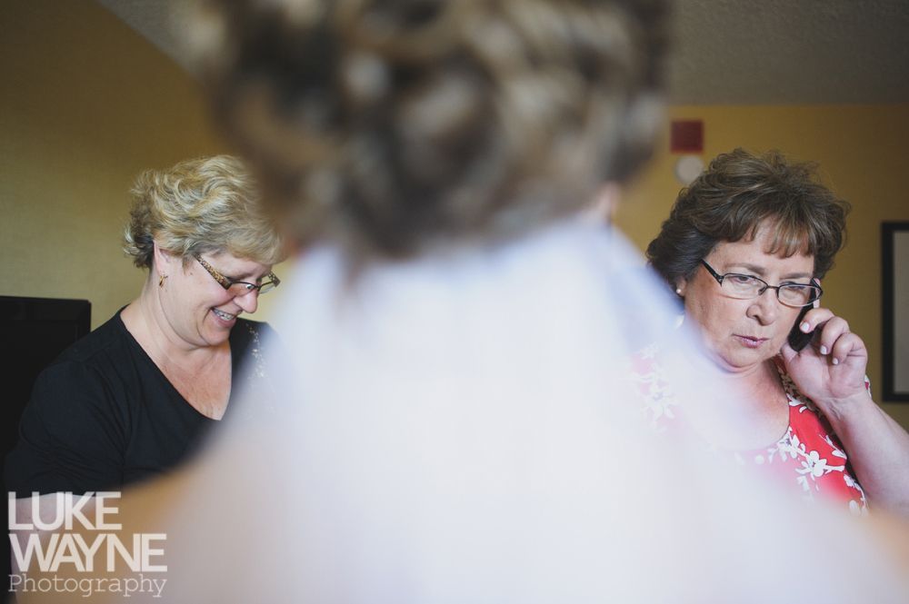 Bride facing camera, blurred. Two women, one smiling, one on phone, observe in a room.