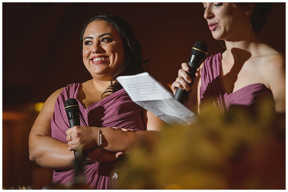 Bride and groom embrace outdoors, veil billowing, wedding bouquets visible.