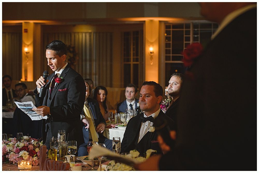 Bride and groom embrace outdoors, veil billowing, wedding bouquets visible.
