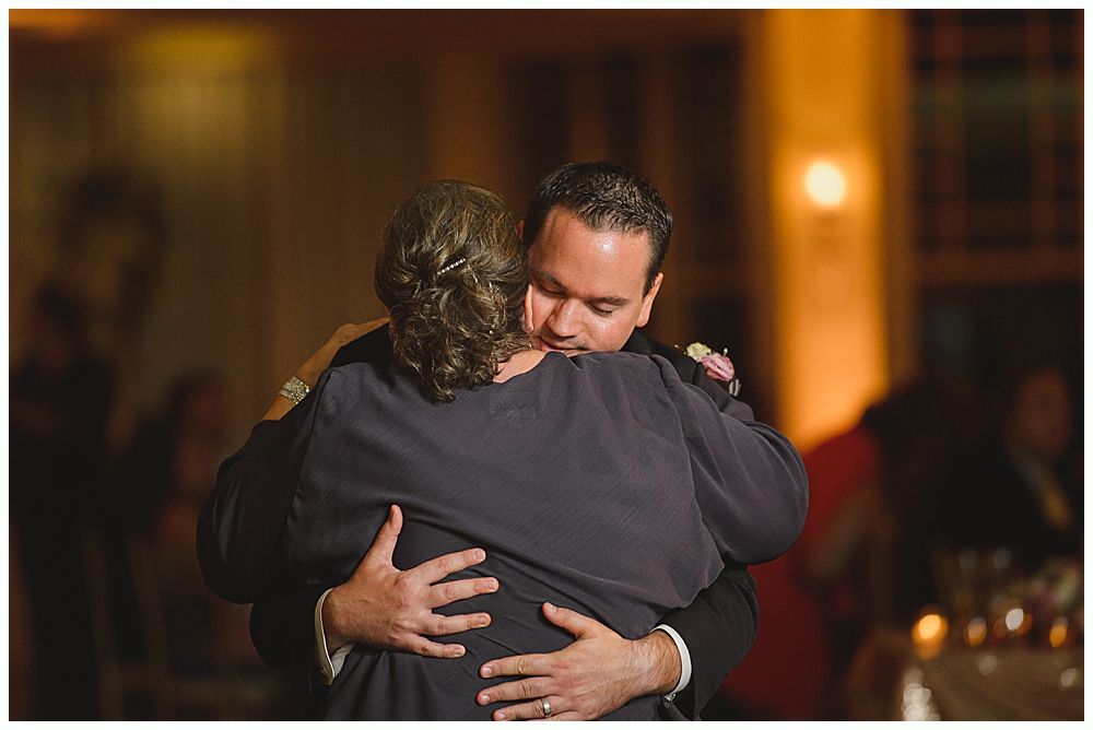 Bride and groom embrace outdoors, veil billowing, wedding bouquets visible.