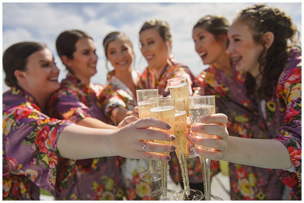 Bridesmaids in red dresses and shawls laugh with bride holding a bouquet outdoors.
