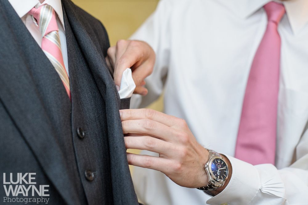 Person adjusting another person's pocket square, both wearing suits and pink ties.